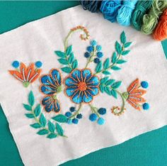 an embroidered piece of cloth with blue flowers and green leaves on it, next to yarn balls