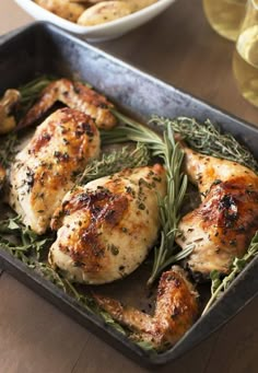 a pan filled with chicken and herbs on top of a wooden table next to wine glasses