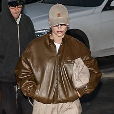 a man in a leather jacket and hat walking down the street with his hand on his hip