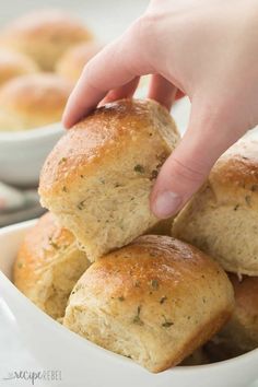 a person picking up some bread from a white bowl with other rolls in the background