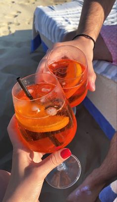 two people holding up wine glasses with drinks in them on the sand at the beach