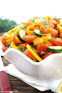 a white dish filled with lots of vegetables on top of a wooden table next to a knife