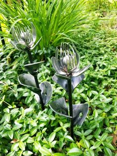 two metal flowers with forks in the middle of some green leaves and plants behind them