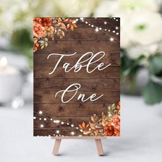 a wooden sign that says table one with flowers and lights on it in front of a candle