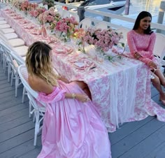 two women sitting at a long table with pink flowers on it and one woman standing in front of the table