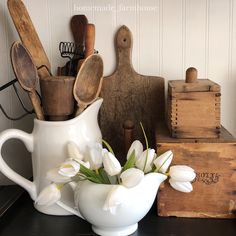 white tulips and wooden spoons sit in a pitcher on a table next to other kitchen utensils