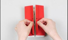 two hands holding an open red book on top of a white wall with one hand