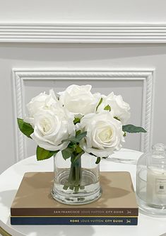 white roses in a glass vase on top of a book next to a candle holder