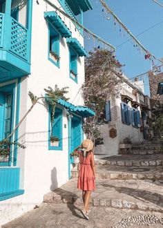 a woman in a red dress and straw hat walks down the street past blue shuttered buildings