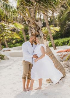 a man and woman standing under a palm tree on the beach with their arms around each other