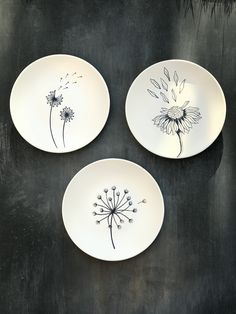 three white plates with dandelions painted on them sitting on a black table top