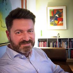 a man with a beard sitting in front of a book shelf and looking at the camera