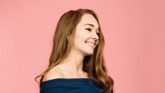 a woman with long brown hair wearing a blue top and smiling at the camera while standing against a pink background