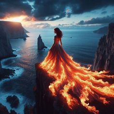 a woman standing on top of a cliff next to the ocean with fire flowing from her dress