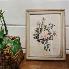 a framed flower arrangement next to a woven basket and potted plant on a wooden table