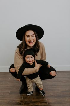 a woman holding a baby wearing a black hat and smiling at the camera with her hands on her chest