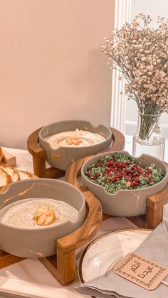 three bowls of food on a table with bread and flowers in vases behind them