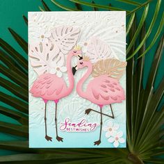 two pink flamingos standing next to each other on top of a palm leaf covered table