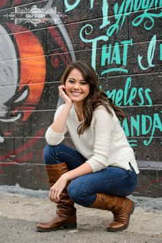 a woman sitting on the ground in front of a wall with graffiti and wearing boots