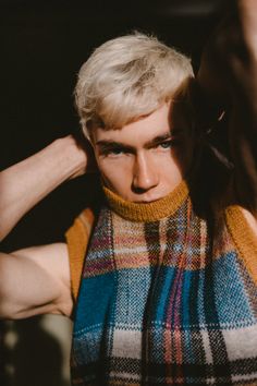 a young man with blonde hair wearing a plaid vest and turtle neck sweater over his shoulders