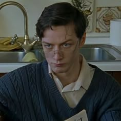 a man with glasses holding a book in his hand and looking at the camera while standing next to a sink