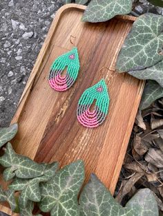 pink and green beaded earrings sitting on top of a wooden board next to leaves