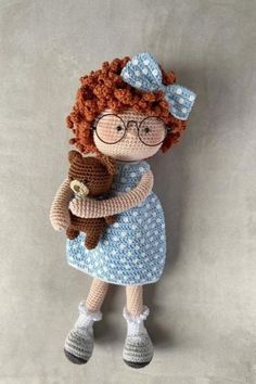 a crocheted doll holding a teddy bear wearing glasses and a blue dress is posed against a gray background