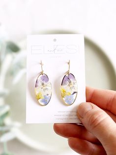 a person holding up a pair of earrings in front of a white plate with flowers on it