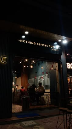 the outside of a coffee shop at night with lights on and people sitting in chairs