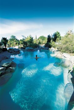 a person swimming in a pool with rocks and trees around it on a sunny day