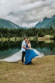 a man and woman standing next to each other in front of a lake surrounded by mountains