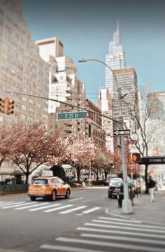 a yellow taxi driving down a street next to tall buildings and traffic lights with cherry blossoms on the trees