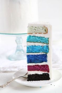 a slice of rainbow cake on a plate with a fork next to it and a glass vase in the background