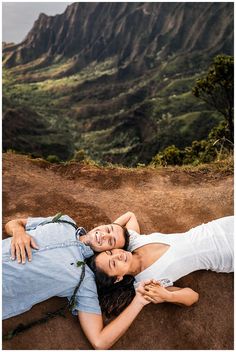 two people are laying on the ground in front of mountains and trees, one person is holding his arm around the woman's shoulder