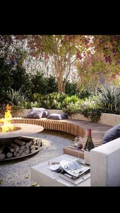 an outdoor fire pit surrounded by trees and rocks with wine glasses on the table next to it