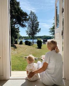 a woman sitting on the ground holding a baby next to an open door with a lake in the background