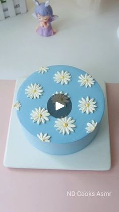 a blue cake with white daisies on it sitting on top of a table next to a figurine