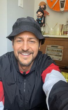 a man wearing a hat and jacket sitting in front of a wall with clocks on it