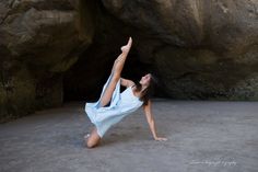 a woman in a blue dress is doing yoga on the beach near a rock formation