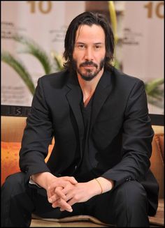 a man in a black suit sitting on a couch with his hands crossed and looking at the camera