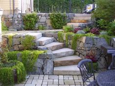 stone steps lead up to a patio with table and chairs in the foreground, surrounded by greenery