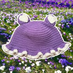 a purple crocheted bear hat laying on the ground in a field of flowers