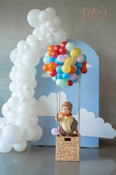 a baby doll sitting in a basket with balloons attached to the handle, and an arch behind it