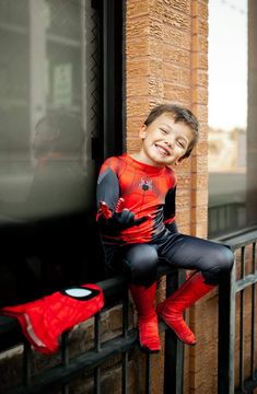 a young boy dressed in a spiderman costume sitting on a railing talking on a cell phone