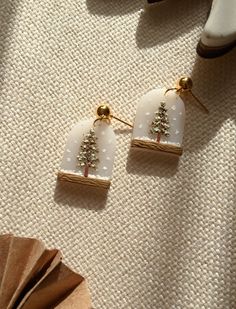 a pair of white and gold earrings with trees on them sitting next to a fan