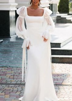 a woman wearing a white wedding dress with long sleeves and bows on her head standing in front of a building