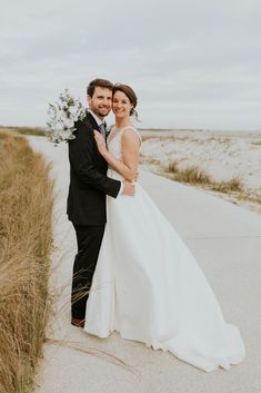 a bride and groom pose for a photo on the beach in front of some tall grass