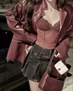 a woman standing next to a car wearing a red leather jacket and skirt with her hands in her pockets