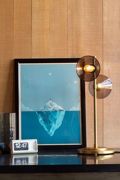 Amplitude Table Lamp sits on a metal desk with a modern flip clock and smoke glass vase. A wood paneled wall is behind the arrangement with a framed picture frame leaning against it. Home Lighting, Modern Lighting, Light Bulbs, Office Space, Table Lamp, California