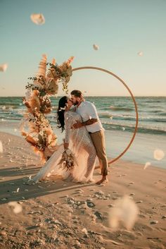 a newly married couple standing on the beach under an arch decorated with flowers and leaves